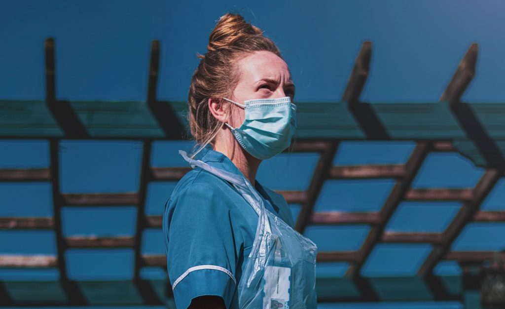 A white woman NHS clinician wears a blue disposable mask and other personal protective equipment.